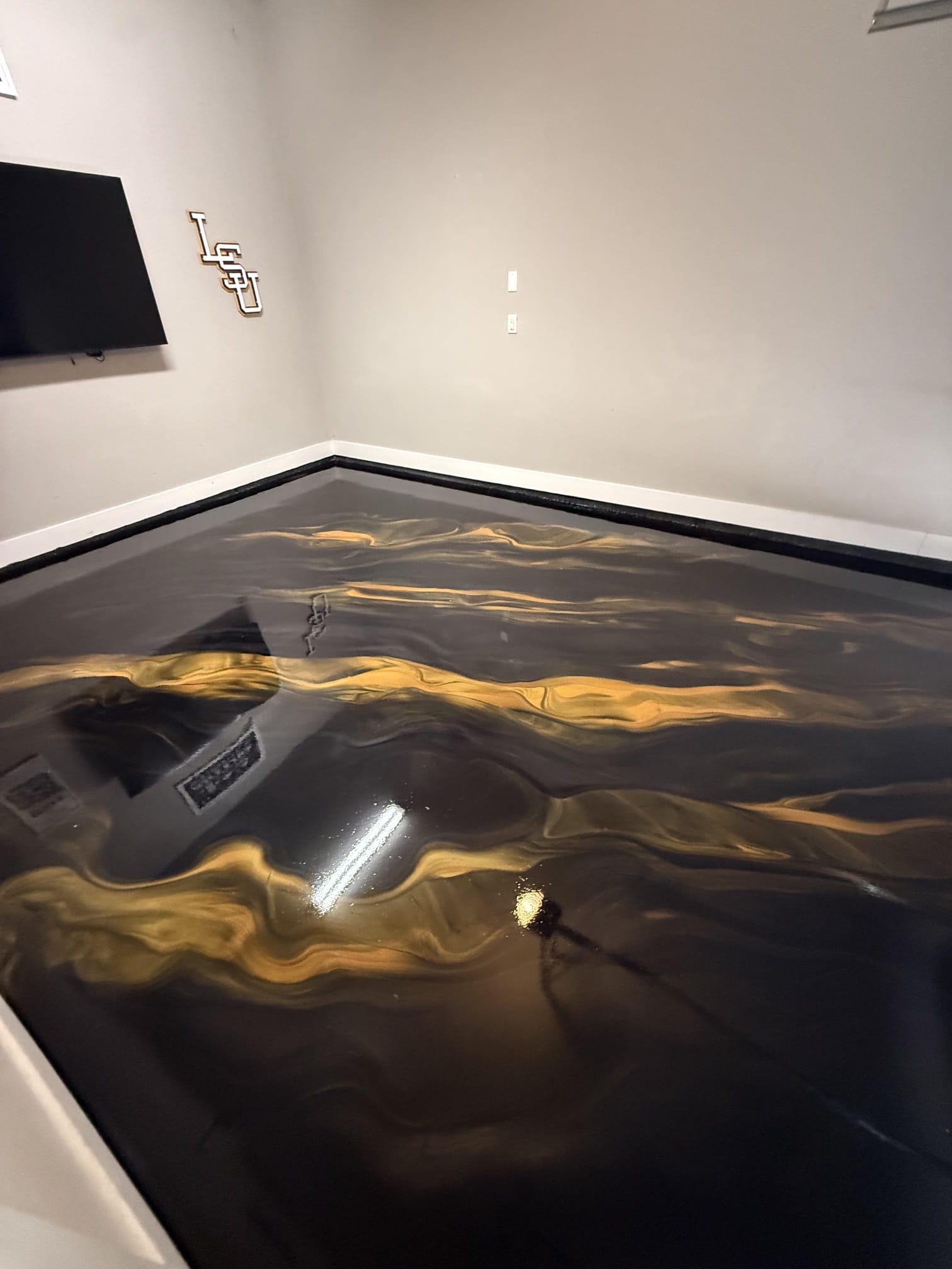 Room with a wall TV and LSU letters on the wall, black and gold metallic epoxy floor with gold ribbons in Shenandoah, LA
