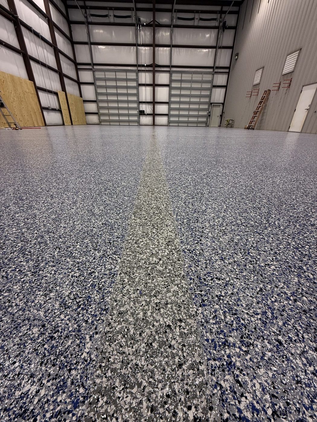Tall metal shop building with roll-up doors, blue, gray, black and white flake floor with a darker flake stripe down the middle, ladders and plywood along the walls in Shenandoah, LA